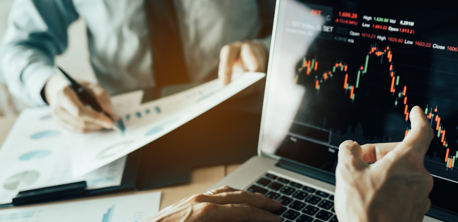 2人の投資家が協力して、紙のストックデータグラフを分析し、ラップトップ画面上のデータを表示しています。